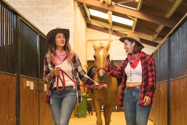 Two cowgirl women having fun and laughing with a horse in a stable, with South American outfits