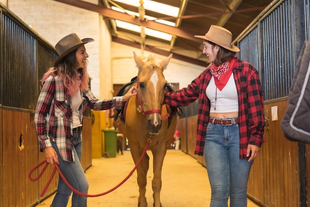 Two cowgirl women having fun and laughing with a horse, with south american outfits, lifestyle of two women working in a stable