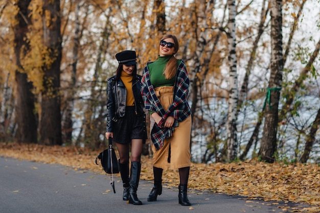 Due ragazze sorridenti accoglienti camminano alla strada del parco di autunno