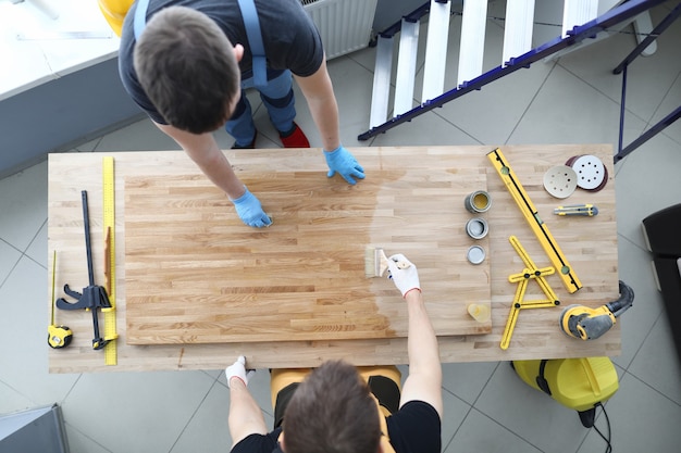 Two craftsmen are covering wooden table with protective varnish