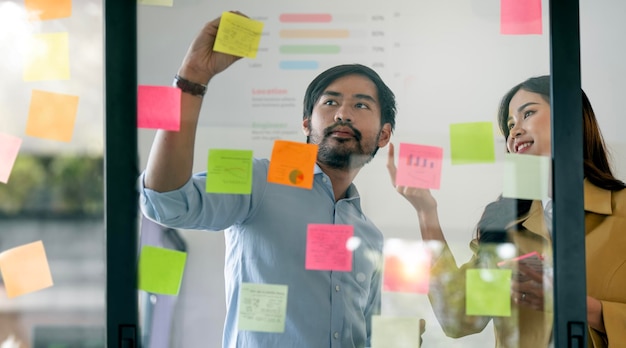 Two Creative business people brainstorming writing on sticky notes on glass wall in office