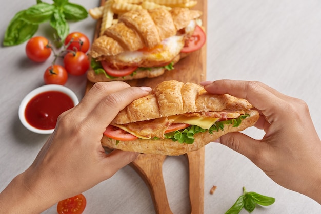 Two croissant sandwiches on wooden table, top view, Sandwich with bacon, fried egg. Ham, cheese, bacon, fried egg, tomato, french fries and lettuce served in hands.