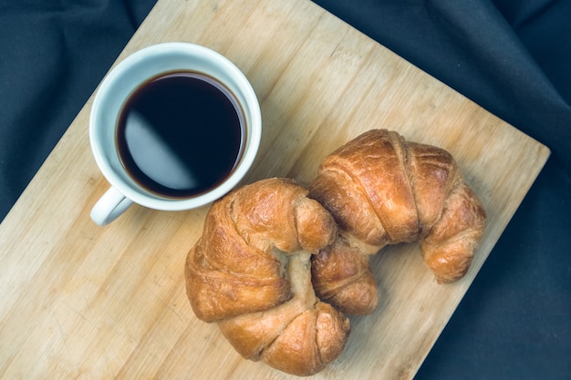 Two croissants with a glass of hot coffee on a chopping board.