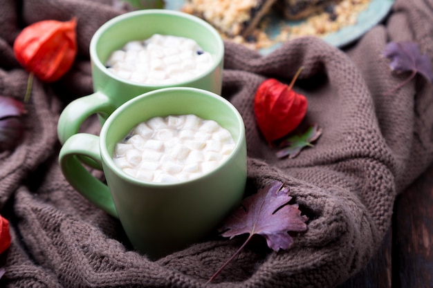 Due tazze di caffè o cioccolata calda con marshmallow vicino a maglia coperta e torta.