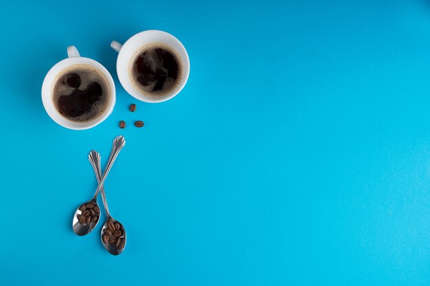 Photo two cups of black coffee, beans and spoons on blue. top view