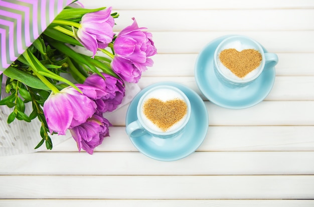 Two cups of cappuccino with a heart shaped symbol and purple tulips on a wooden background