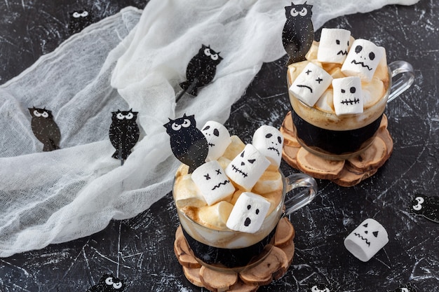 Two cups coffee with marshmallow (spooky faces, monster) on dark table for Halloween