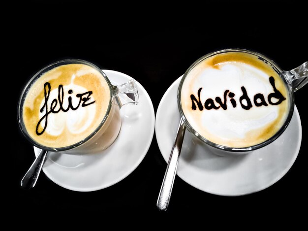 Two cups of coffee with milk with latte art lettering Merry Christmas, on black background.