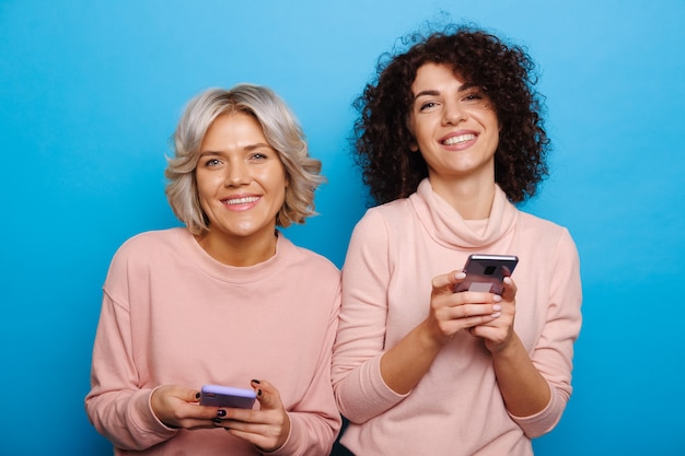 Two curly haired caucasian women are chatting and smiling at camera on a blue wall