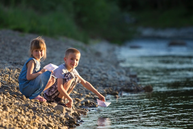 Due bambini biondi svegli, ragazzo e ragazza sulla sponda del fiume che inviano le barche del libro bianco dell'acqua