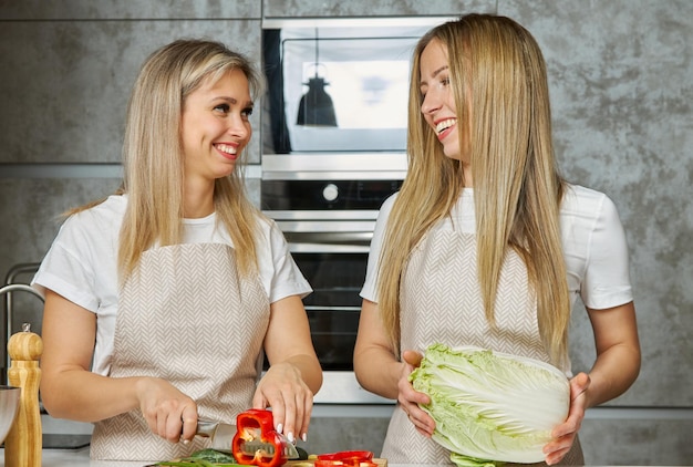 Two cute blonde women cook together from fresh vegetables and\
smile gently at each other in a modern kitchen