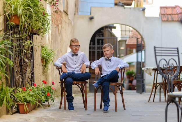 Две милые мальчики разговаривают сидя на деревянных стульях. мальчики подражают родителям бизнесменам. мальчики сидят на стульях со скрещенными ногами.