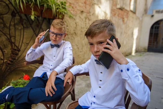 Due ragazzi carini stanno parlando sul loro smartphone. i ragazzi imitano i genitori agli uomini d'affari. i ragazzi sono seduti su sedie di legno sulla strada della città vecchia.