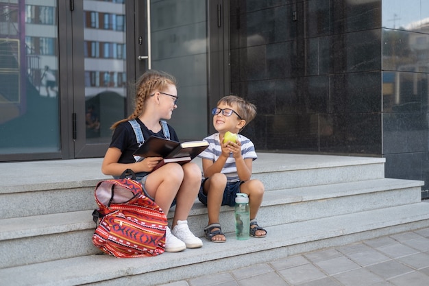 책과 배낭이 달린 안경을 쓴 두 명의 귀여운 백인 행복한 아이들. 앉아서 점심을 먹습니다. 방학. 지식의 날. 학교 개념으로 돌아가기.