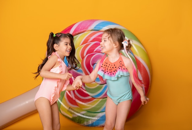 Two cute child girls in swimming suit laughing having fun on yellow surface with lollipop.