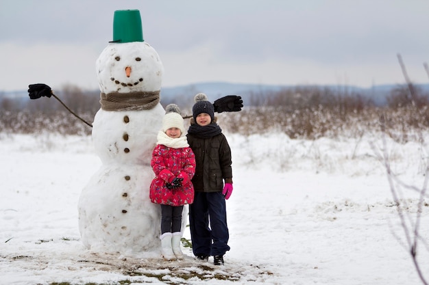 Two cute children, boy and girl, standing in front of smiling snowman in bucket hat, scarf and gloves on snowy winter landscape and blue sky copy space background. Merry Christmas, Happy New Year.