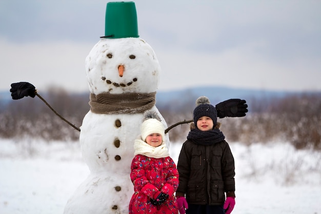 Two cute children, boy and girl, standing in front of smiling snowman in bucket hat, scarf and gloves on snowy winter landscape and blue sky copy space background. Merry Christmas, Happy New Year.