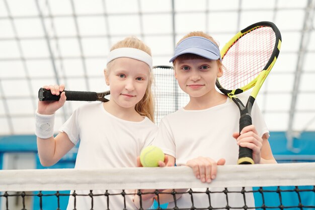 Two cute friendly girls in white sportswear standing by net at stadium in front of camera and holding tennis rackets on shoulders ready for play