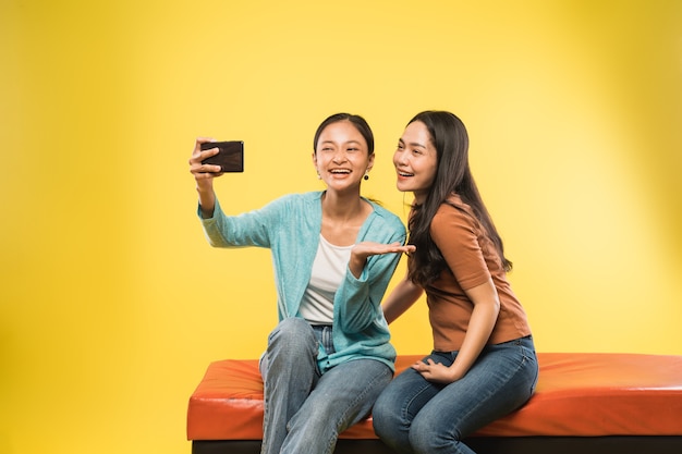 Две милые девушки с помощью камеры смартфона, чтобы сделать селфи вместе, сидя