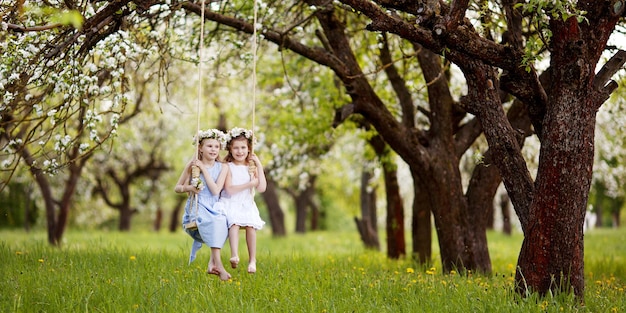 Two cute girls having fun on a swing in blossoming old apple tree garden Sunny day Spring outdoor activities for kids