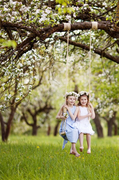 Two cute girls having fun on a swing in blossoming old apple tree garden Sunny day Spring outdoor activities for kids