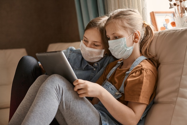 Two cute girls in medical masks are sitting on sofa with a tablet.