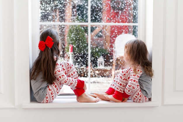 Two cute girls in pajamas sitting and looking out the window at snowy weather.
