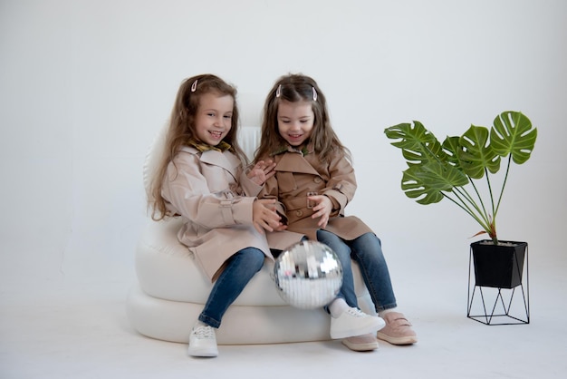 Two cute little girls in identical trench coats sits in a white chair White background Fashion