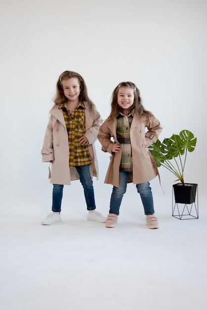 Two cute little girls in identical trench coats stand together White background Fashion Childhood