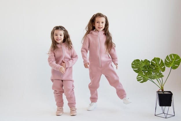 Photo two cute little girls in a pink overalls sits stand together white background fashion childhood