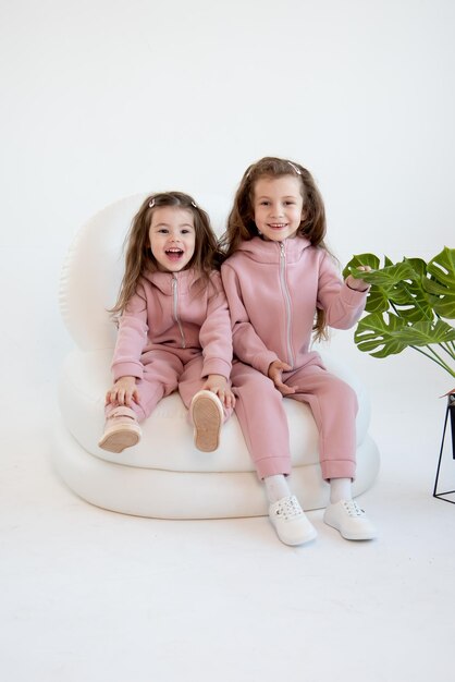 Two cute little girls in a pink overalls sits in a white chair White background Fashion