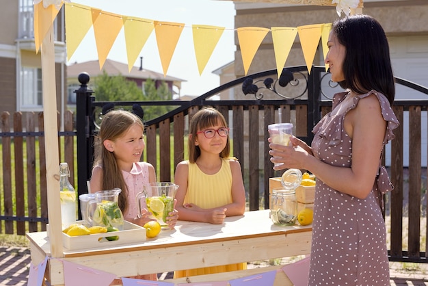 Two cute little girls standing by wooden stall and looking at young pretty female in elegant dress having glass of fresh homemade lemonade