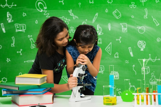 Две милые маленькие индийские или азиатские школьницы, экспериментирующие или изучающие науку в лаборатории, на фоне зеленой классной доски с образовательными рисунками