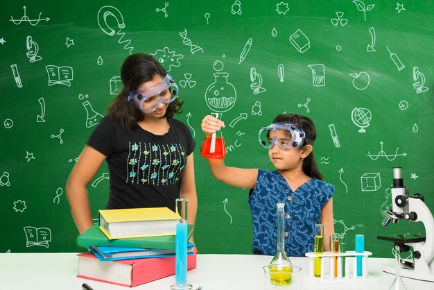 実験室で科学を実験または勉強している2人のかわいい小さなインド人またはアジア人の女子学生、教育的な落書きで緑の黒板の背景の上に
