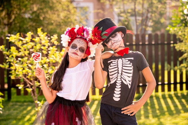 Two cute little kids with painted faces standing close to one another in front of camera and looking at you while celebrating halloween