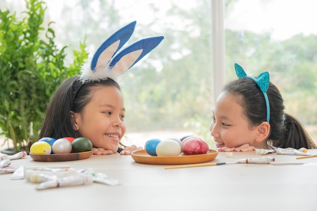 Photo two cute little sisters wearing bunny ears playing egg hunt on easter adorable children celebrate easter at home teenage asian girl painting and decorating easter eggs