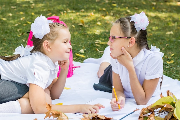 Two cute schoolgirls are smiling and doing homework on a blanket in a sunny autumn park. Outdoor education. Back to school concept