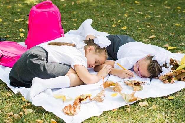 Two cute schoolgirls were tired of doing their homework and fell asleep on a blanket in a park