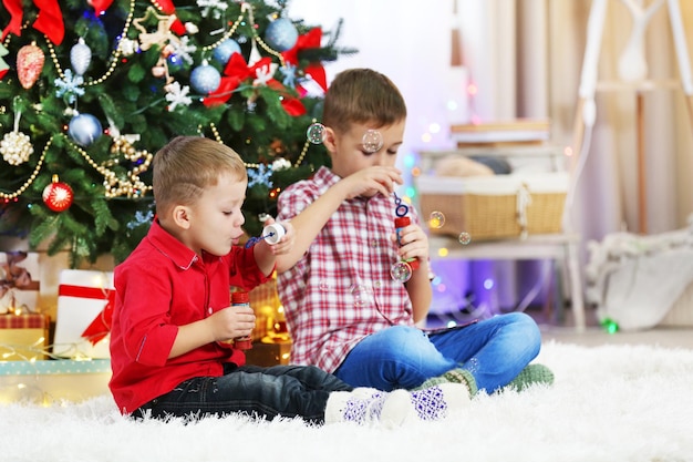 Two cute small brothers blows soap bubbles on Christmas tree background