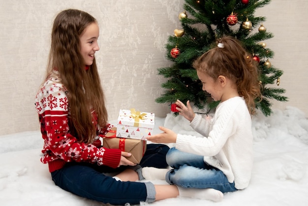 Two cute smiling girls sit next to the Christmas tree and give each other gifts New Year Christmas