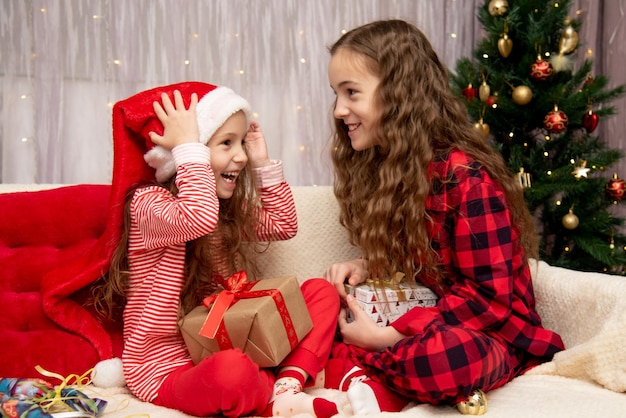 Two cute smiling girls sit next to the Christmas tree, have a fan, and give each other gifts.