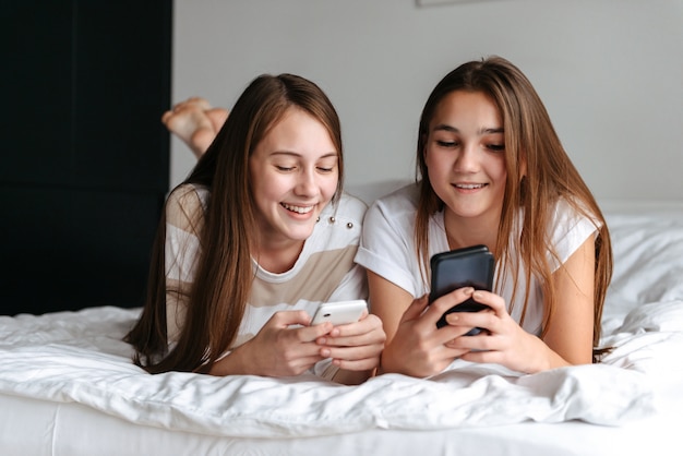 Two cute smiling teengae girls laying on bed, holding mobile phones at home