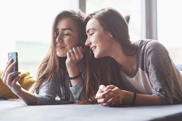 Two cute smiling twins sisters holding smartphone and making selfie. Girls lie on the couch posing and joy