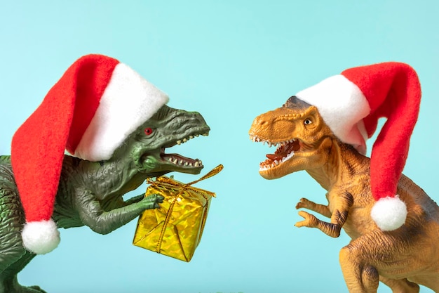 Two dinosaurs rex in red santa claus hat holds golden gift box\
in its paws on blue background