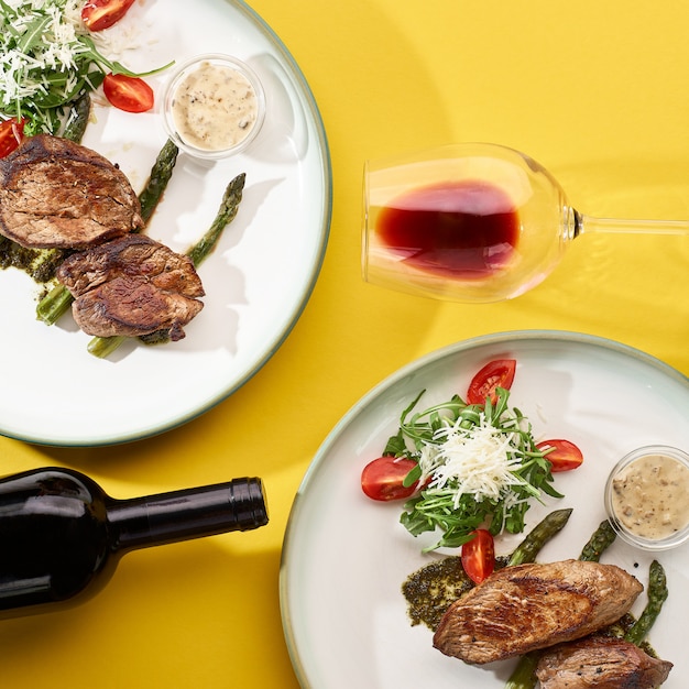 Two dishes with grilled pork meat with fresh vegetables salad and red wine on a yellow wall. top view