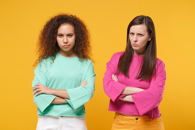 Due donne dispiaciute amiche ragazze europee e afroamericane in abiti rosa verdi in posa isolati su sfondo giallo arancione. concetto di stile di vita delle persone. mock up spazio di copia. tenendosi per mano incrociata.