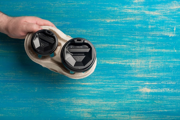 Two disposable paper cups of coffee and a cup holder in a male hand on a blue wooden surface