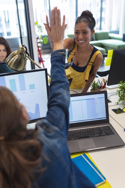 Photo two diverse female creative colleagues sitting at desk high-fiving and smiling. modern office of a creative design business.