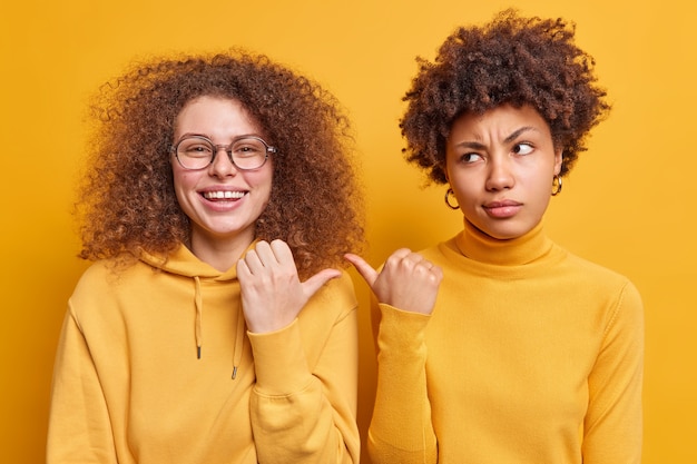 写真 黄色い壁に孤立した幸福と不満をさりげなく表現する服を着た2人の多様なフレンドリーな女性がお互いに親指を向けます。私の友達を見てください。屋内の混血の女性の友人