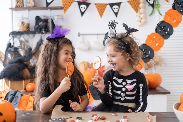 Due ragazze diverse in costume da strega, si divertono in cucina, mangiano biscotti, festeggiano halloween.